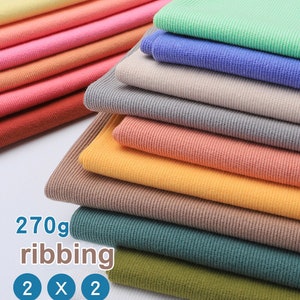Puede incluir: Una pila de tejido de punto de canalé de 270 g en varios colores, incluidos amarillo, verde, azul, gris, naranja, marrón, verde azulado y blanco. El tejido está dispuesto en forma escalonada, con el texto "270g ribbing 2 x 2" visible en la parte inferior de la imagen.