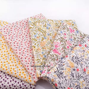 May include: Several fabric samples with floral patterns. The designs include small flowers and foliage in pink, yellow, blue, and purple, set on a white background. The website address fabricmade.etsy.com is visible on the fabric.