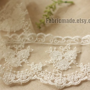 May include: Ivory lace trim with floral and scrollwork embroidery. The delicate fabric features a scalloped edge and is suitable for various craft projects. The text "Fabricmade.etsy.com" is visible.