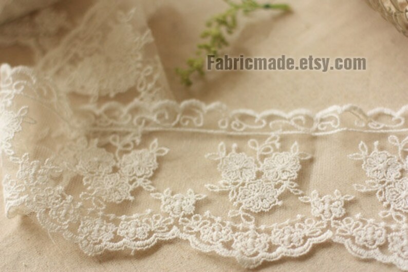 May include: Ivory lace trim with floral and scrollwork embroidery. The delicate fabric features a scalloped edge and is suitable for various craft projects. The text "Fabricmade.etsy.com" is visible.