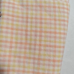 May include: Close-up of a fabric with a gingham pattern. The fabric features a repeating grid of squares in shades of peach, yellow, and white. The fabric appears to be soft and lightweight, suitable for clothing or home decor.