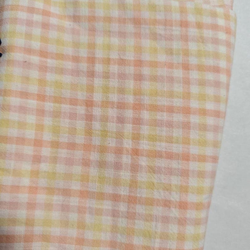 May include: Close-up of a fabric with a gingham pattern. The fabric features a repeating grid of squares in shades of peach, yellow, and white. The fabric appears to be soft and lightweight, suitable for clothing or home decor.