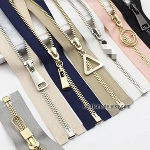 Pode incluir: Variedade de zíperes em várias cores, incluindo cinza, bege, azul marinho e rosa. Os zíperes têm diferentes designs de puxadores de metal, como um triângulo, um rosto sorridente e uma forma retangular. O site Fabricmade.etsy.com é visível.