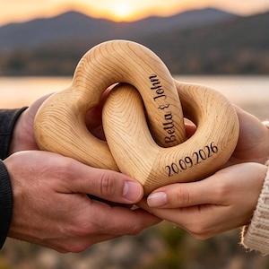 May include: Two interlocking light-colored wooden heart shapes with a visible wood grain texture. The surface is engraved with the text Bella & John and the date 20.09.2026 in a dark brown script and sans-serif font.