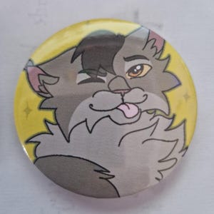 Pu&ograve; includere: Una spilla rotonda con l'illustrazione a cartone animato di un gatto grigio e soffice con una macchia nera sulla testa. Il gatto fa l'occhiolino e tira fuori la lingua. Lo sfondo &egrave; un cerchio giallo brillante con piccoli dettagli a forma di stella.