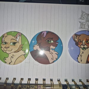 May include: Three circular pin-back buttons featuring stylized illustrations of cats. The left button has a light tan cat on a green background. The center button shows a brown cat on a blue background. The right button depicts a calico cat on a purple background.