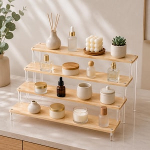 May include: A four-tiered display stand featuring light wood shelves supported by clear acrylic pillars. The shelves hold various items including glass perfume bottles, small ceramic jars, a bubble candle, a succulent, and a reed diffuser.