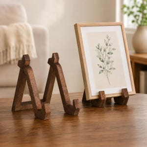 Könnte beinhalten: Ein dunkelbrauner Holzständer mit dreieckigem Rahmen und einer eingekerbten Ablagefläche. Daneben befindet sich ein heller Holzbilderrahmen mit einem Druck, der eine botanische Illustration grüner Blätter auf cremefarbenem Grund zeigt.