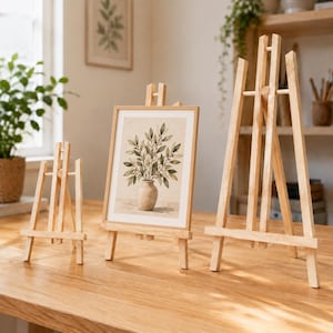 Può includere: Tre cavalletti in legno di colore chiaro di diverse altezze disposti su una superficie in legno. Il cavalletto centrale sostiene una stampa rettangolare incorniciata con un'illustrazione botanica di una pianta a foglie in un vaso.