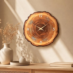 May include: A round wall clock crafted from a natural wood slice with visible dark brown tree rings and a live edge. The clock face features white numerals at the 12, 3, 6, and 9 positions, small white dot markers for other hours, and thin gold hands.
