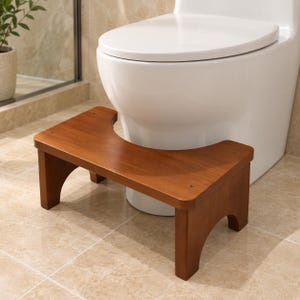 May include: A brown wooden toilet stool with a curved cutout design, featuring a smooth rectangular top surface and four sturdy legs, positioned directly in front of a white porcelain toilet base on a tiled bathroom floor.
