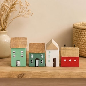 Può includere: Un set di quattro piccole statuette di case in legno con tetti color legno naturale. Le case sono dipinte in verde chiaro, verde scuro, bianco e rosso, con finestre, porte e piccoli dettagli dipinti a mano sulle facciate anteriori.