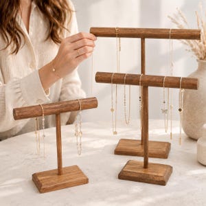May include: Two dark wood T-shaped jewelry stands with rectangular bases. The stands hold multiple gold-toned necklaces with delicate chains, small circular pendants, and a butterfly charm. The wood has a visible grain and smooth finish.