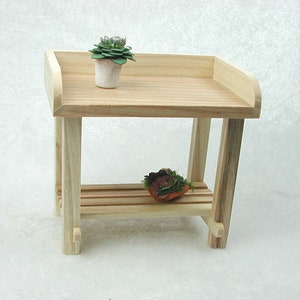 May include: A wooden two-tiered shelf with a natural finish. The top shelf has a rounded edge and the bottom shelf has a slatted design. There are two small potted plants on the shelves.