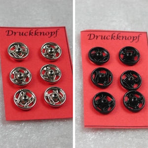 Puede incluir: Dos tarjetas con la palabra "Druckknopf" impresa en ellas. Cada tarjeta tiene seis botones a presión de metal, tres plateados y tres negros.