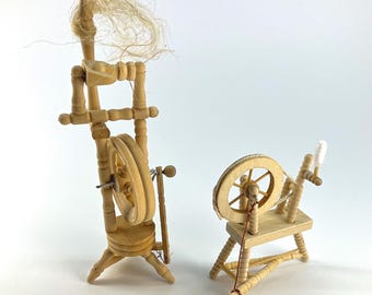 May include: Two miniature wooden spinning wheels with light tan natural wood tones. The taller vertical model features a distaff with loose flax fibers at the top, while the smaller horizontal model includes a circular wheel and a white fiber bobbin.