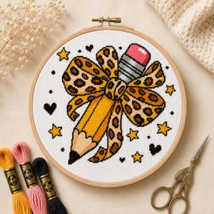 May include: A cross-stitch project in a round wooden embroidery hoop featuring a yellow pencil with a pink eraser and a large leopard print bow. The design is surrounded by small black hearts and yellow stars on white fabric, with embroidery floss.