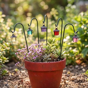 Könnte beinhalten: Ein Terrakotta-Blumentopf mit verschiedenen grünen Sukkulenten und kleinen violetten Blüten, ausgestattet mit fünf schwarzen Metall-Hirtenstäben. Jeder Stab trägt eine Miniatur-Laterne in transparent, türkis, violett, rot und rosa.
