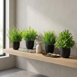 May include: Four artificial green plants in matte black pots arranged on a wooden shelf. Three plants have broad leaves, one has thin grass-like blades. A white ceramic diffuser sits between the plants, and a small gold tray holds eyeglasses.