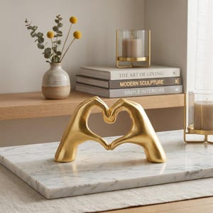 May include: A gold-colored sculpture depicting two hands forming a heart shape. The sculpture has a smooth, matte finish and is positioned on a white marble surface. The hands are joined at the fingertips and thumbs to create a central heart-shaped opening.