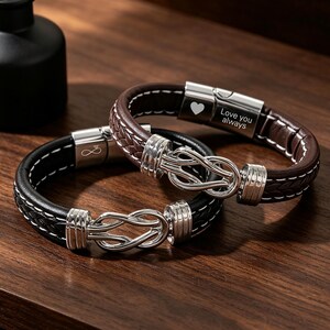 May include: Two leather bracelets with silver-toned metal clasps. One bracelet is black with white stitching and an infinity symbol on the clasp. The other is brown with white stitching, a heart icon, and the text "Love you always" on the clasp.