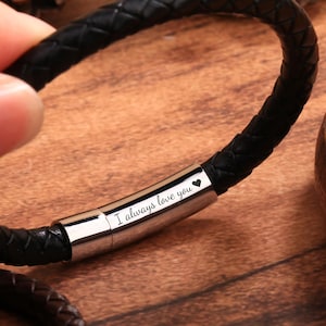 May include: A black braided leather bracelet featuring a polished silver-tone metal cylindrical clasp. The clasp is engraved with the text "I always love you" followed by a small heart symbol. The bracelet has a smooth, woven texture.
