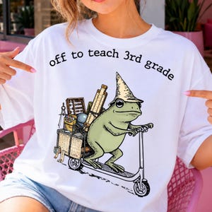 May include: A white t-shirt featuring a graphic of a green frog wearing a pointed hat and riding a scooter. The frog carries school supplies including an abacus, a ruler, and a compass. The text "off to teach 3rd grade" is printed above.