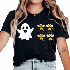 May include: A dark charcoal gray short-sleeve t-shirt featuring a graphic print on the front. The design includes a single white cartoon ghost with a smiling face positioned to the left of four small yellow and black cartoon bees.