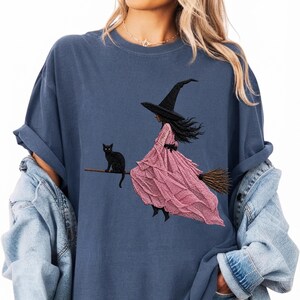Puede incluir: Una camiseta azul pizarra con un estampado gr&aacute;fico de una figura con sombrero puntiagudo negro y un vestido largo rosa texturizado montada en una escoba. Un gato negro est&aacute; sentado en la escoba detr&aacute;s de la figura.