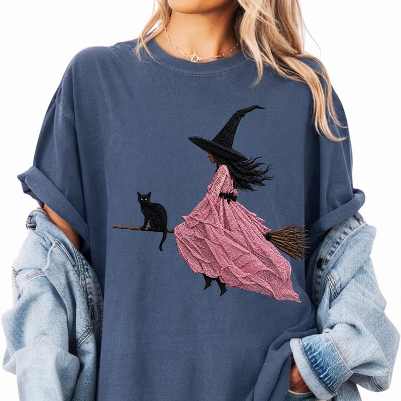 Puede incluir: Una camiseta azul pizarra con un estampado gr&aacute;fico de una figura con sombrero puntiagudo negro y un vestido largo rosa texturizado montada en una escoba. Un gato negro est&aacute; sentado en la escoba detr&aacute;s de la figura.