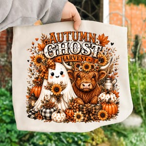 Peut inclure: Un sac fourre-tout couleur crème avec une impression graphique représentant un fantôme et une vache highland entourés de tournesols, de citrouilles et de feuilles d'automne. Le texte indique AUTUMN GHOST HARVEST. Le motif inclut des carreaux.
