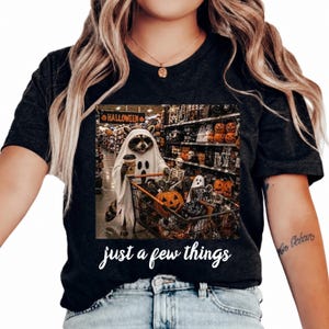 Peut inclure: Un t-shirt noir avec une illustration représentant un raton laveur déguisé en fantôme, poussant un caddie rempli de décorations d'Halloween dans un magasin. Le texte "just a few things" est imprimé en écriture blanche sous l'image.