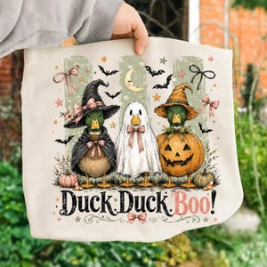 Peut inclure: Un sac fourre-tout crème avec une illustration de trois canards déguisés pour Halloween : un en sorcière, un en fantôme et un dans une citrouille. Le motif inclut des chauves-souris, des étoiles et des nœuds, avec le texte Duck Duck Boo!