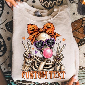 Puede incluir: Una sudadera beige de cuello redondo con el gráfico de un esqueleto que lleva gafas de sol moradas en forma de corazón con telarañas, un lazo naranja con caras de calabaza y haciendo una burbuja rosa. El texto CUSTOM TEXT aparece debajo.