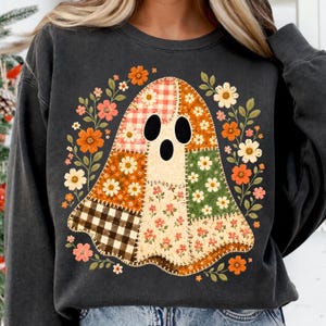 Puede incluir: Una sudadera de cuello redondo gris carbón con un gráfico central de fantasma compuesto por varios patrones de patchwork, incluyendo cuadros vichy rojos y blancos, cuadros marrones y blancos, y estampados florales en naranja, verde y crema, rodeado de flores.