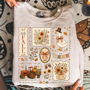 May include: A beige crewneck sweatshirt featuring a graphic print of autumn-themed illustrations including a goose wearing a hat, a tractor carrying pumpkins, floral arrangements, a plaid bow, and the text hello FALL in a vertical orientation.