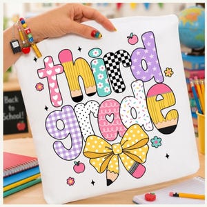 Puede incluir: Un estuche de lona blanca con el texto "third grade" en letras de burbuja coloridas y estampadas. El diseño incluye motivos de lápices, un gran lazo amarillo de lunares, pequeñas ilustraciones de manzanas y detalles florales.