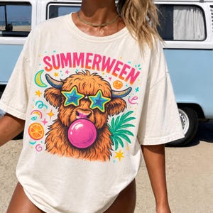 May include: An off-white t-shirt featuring a graphic of a shaggy highland cow wearing star-shaped sunglasses and blowing a pink bubblegum bubble. The text "SUMMERWEEN" is printed in a vibrant pink and orange gradient above the cow illustration.