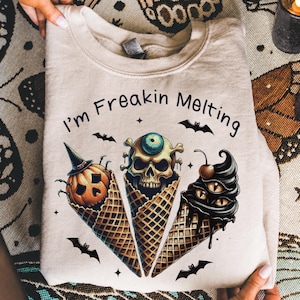 Peut inclure: Un sweat-shirt beige à col rond avec le texte "I'm Freakin Melting" au-dessus de trois cornets de glace gaufrés. Les cornets contiennent une citrouille d'Halloween avec un chapeau de sorcière, un crâne avec un œil et un tourbillon sombre.