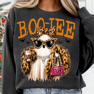 Peut inclure: Un sweat-shirt gris anthracite avec un motif de fantôme portant des lunettes de soleil à imprimé léopard, un nœud assorti et une étole en fausse fourrure. Le fantôme tient un gobelet et un sac à main rose léopard. Le texte BOO-JEE est en orange.