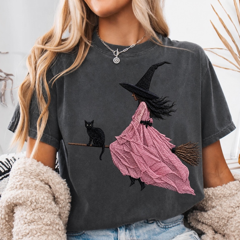 Puede incluir: Una camiseta de manga corta gris carb&oacute;n con un estampado gr&aacute;fico de una bruja con sombrero negro y un vestido largo rosa con volantes, montada en una escoba con un gato negro sentado en el mango.