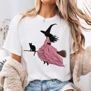 Puede incluir: Una camiseta blanca de manga corta con un estampado gr&aacute;fico de una figura con sombrero negro puntiagudo y un vestido largo de color rosa texturizado, montada en una escoba de madera. Un peque&ntilde;o gato negro est&aacute; sobre la escoba.