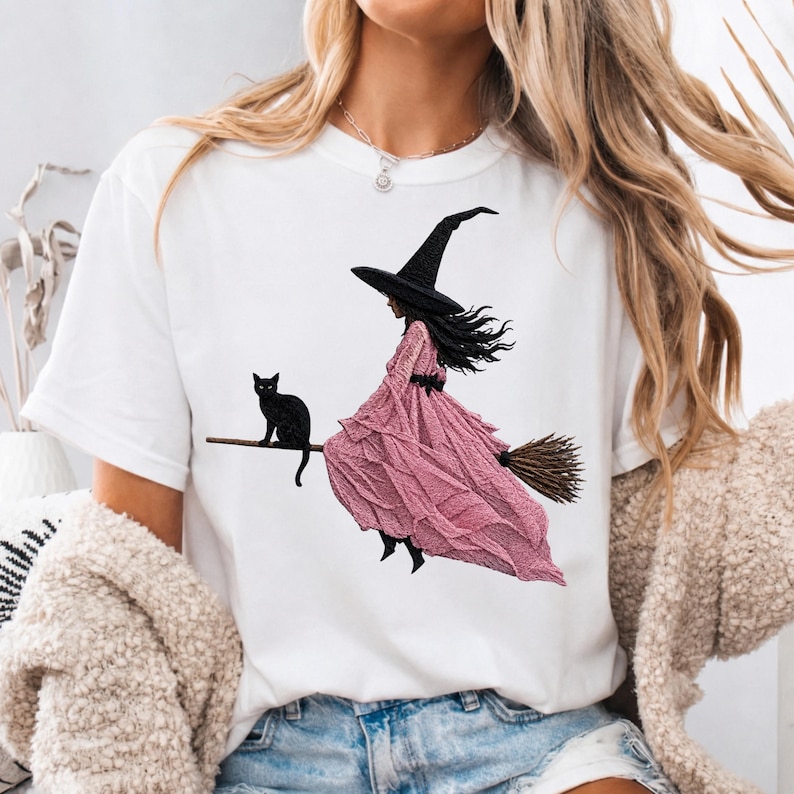 Puede incluir: Una camiseta blanca de manga corta con un estampado gr&aacute;fico de una figura con sombrero negro puntiagudo y un vestido largo de color rosa texturizado, montada en una escoba de madera. Un peque&ntilde;o gato negro est&aacute; sobre la escoba.