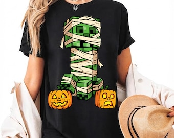 Puede incluir: Una camiseta negra de manga corta con un estampado gráfico de una criatura verde con forma de bloque envuelta en vendas blancas como una momia, situada entre dos calabazas naranjas de Halloween con caras talladas.