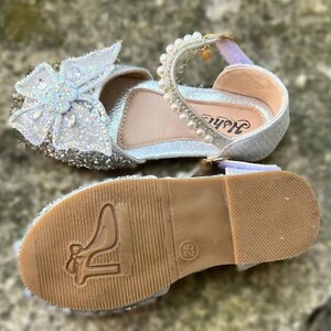 May include: A silver glitter shoe with a large white butterfly ornament on the toe, featuring a strap adorned with white pearls. The tan sole displays a high-heeled shoe icon and the number 25, with a textured grip pattern on the heel area.