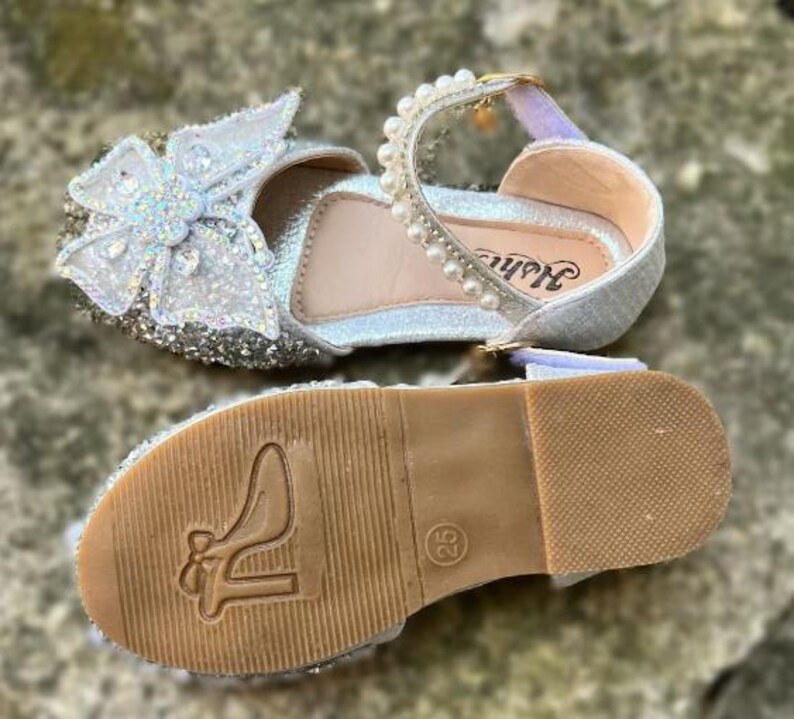 May include: A silver glitter shoe with a large white butterfly ornament on the toe, featuring a strap adorned with white pearls. The tan sole displays a high-heeled shoe icon and the number 25, with a textured grip pattern on the heel area.