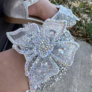 May include: A silver glitter shoe featuring a large, ornate, white mesh bow embellished with iridescent crystals and a central flower motif. The shoe includes a strap across the top adorned with a row of white faux pearls and a flat brown sole.
