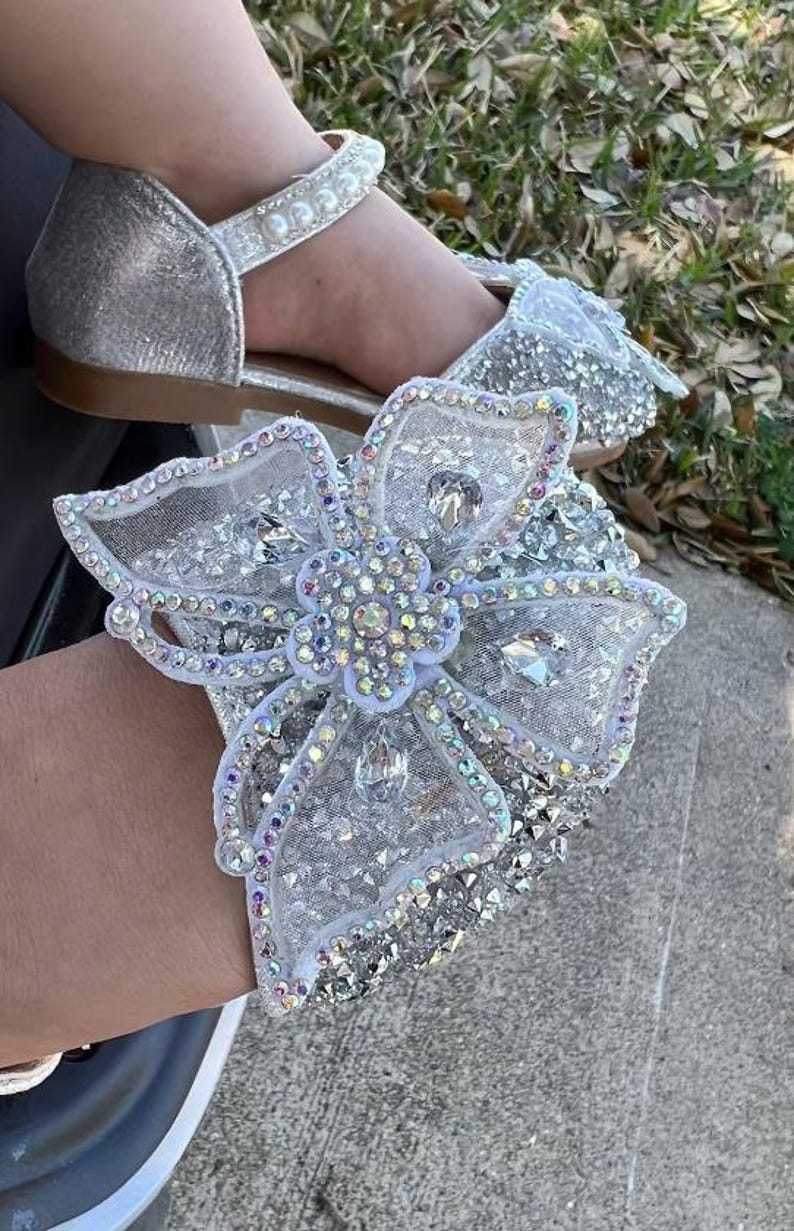 May include: A silver glitter shoe featuring a large, ornate, white mesh bow embellished with iridescent crystals and a central flower motif. The shoe includes a strap across the top adorned with a row of white faux pearls and a flat brown sole.