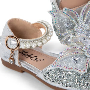 May include: A silver glitter shoe featuring a large butterfly ornament on the toe covered in iridescent crystals. The ankle strap is adorned with a row of white pearls and a gold-toned buckle with a small dangling crystal charm. The brand name Ashtx is printed on the inner sole.