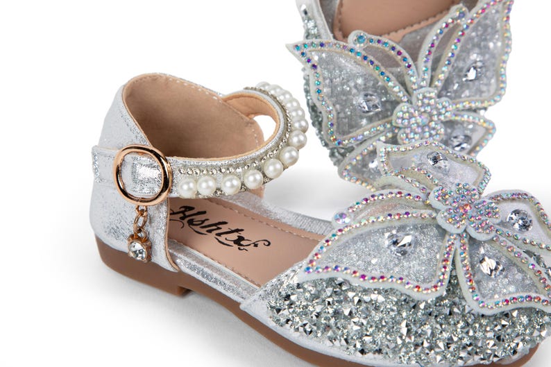 May include: A silver glitter shoe featuring a large butterfly ornament on the toe covered in iridescent crystals. The ankle strap is adorned with a row of white pearls and a gold-toned buckle with a small dangling crystal charm. The brand name Ashtx is printed on the inner sole.
